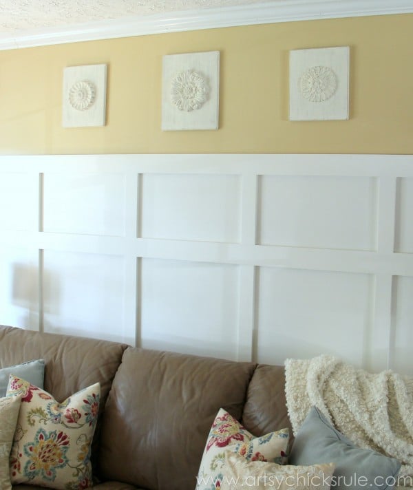 A beige wall features three white wood medallions as decorative panels above white board and batten wainscoting. A tan couch with floral pillows and a textured blanket sits in front.