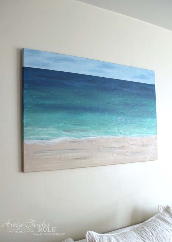 A DIY Beach Painting of a serene beach scene hangs on the wall, depicting sand, turquoise water, and a blue sky. The painting is displayed above a white couch.