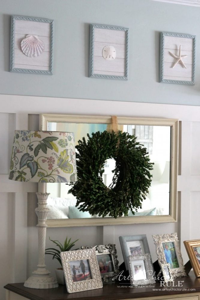 A decorative console table with framed photos, a floral-patterned lamp, and a green wreath hanging on a mirror. Above, three framed seashell wall art pieces—crafted from old prints repurposed—adorn the light blue wall.