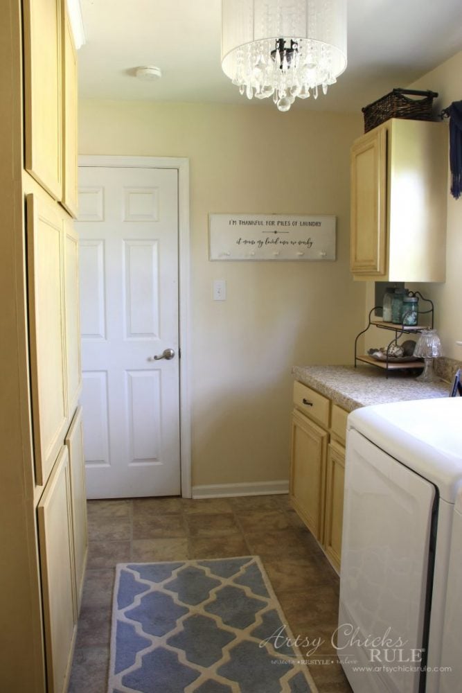 A bright, organized laundry room with light wood cabinets, a washing machine, a patterned rug, and a modern chandelier. Neat and inviting with beige walls and tile flooring, it&rsquo;s perfect inspiration for farmhouse laundry plans.