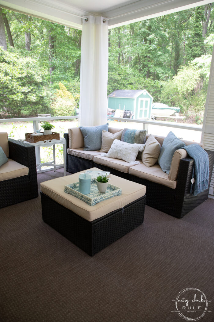 A cozy screened-in porch refresh featuring wicker patio furniture, beige cushions, blue and white throw pillows, a matching ottoman, side table with decor, and views of lush green trees outside.