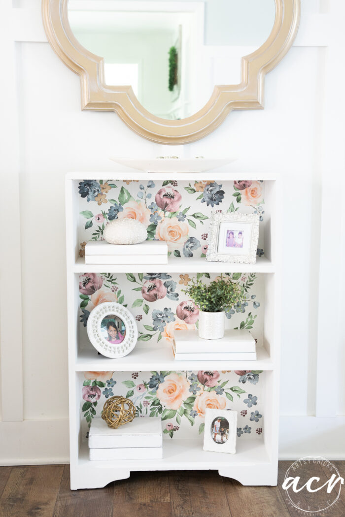 white bookcase with floral backing, gold mirror, styled shelves