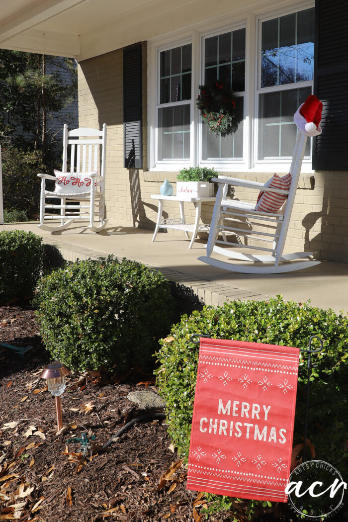 white rockers on porch with small red Merry Christmas flag in flower bed