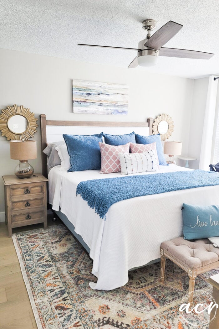 full bedroom shot with blue and white bedding, pillows, colorful rug and ceiling fan
