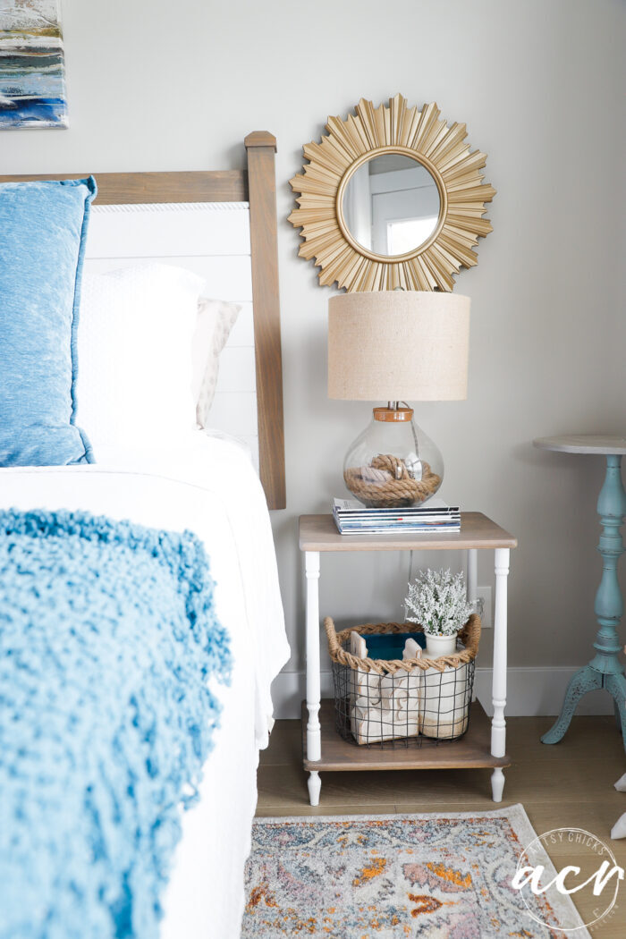 tan and white table with lamp and magazines beside bed with blue and white covers