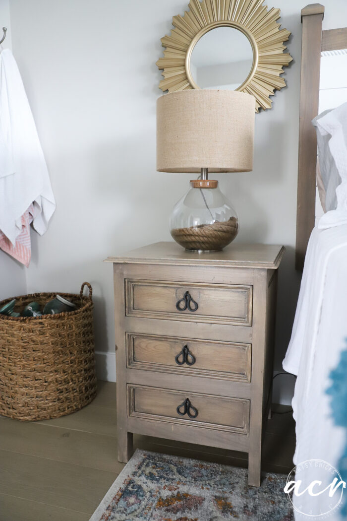 tan nightstand table with lamp beside bed, gold starburst mirror on wall behind