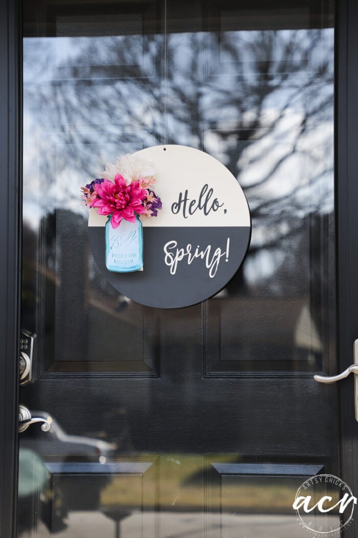 black and white round sign with hello spring, blue jar and pink colorful flowers, on glass front door