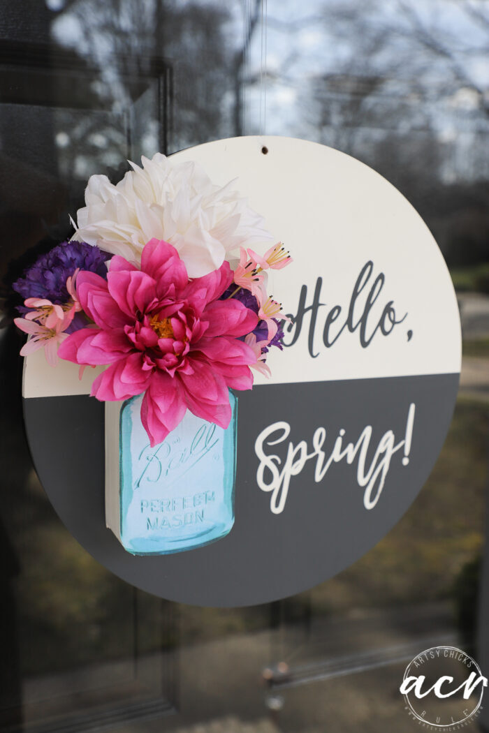 black and white round sign with hello spring, blue jar and pink colorful flowers, on glass front door up close