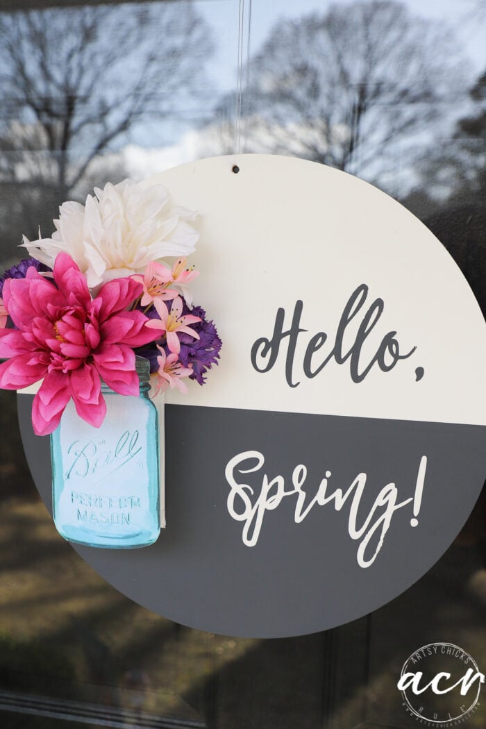 black and white round sign with hello spring, blue jar and pink colorful flowers, on glass front door up close