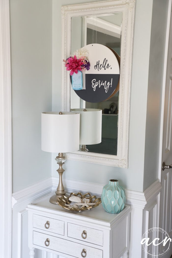 black and white round sign with hello spring, blue jar and pink colorful flowers, on tall white mirror with white cabinet and lamp below