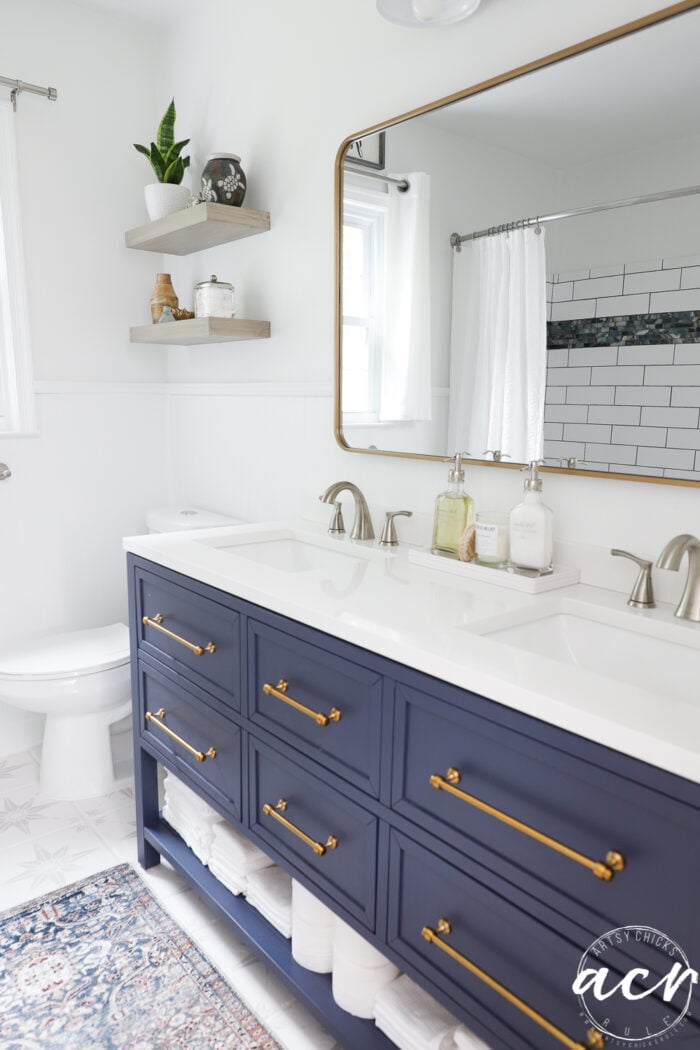 dark blue bath vanity, gold pulls, blue multi color rug, gold mirror, white solid surface top