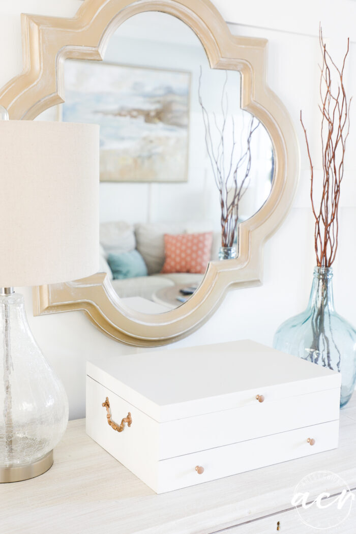 gold mirror on wall, lamp, white box and blue clear glass jar on dresser