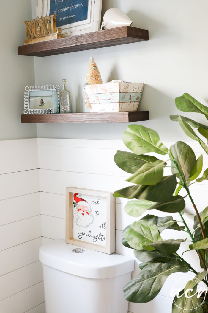 santa sign in bathroom on back of toilet with green plant and shelves above