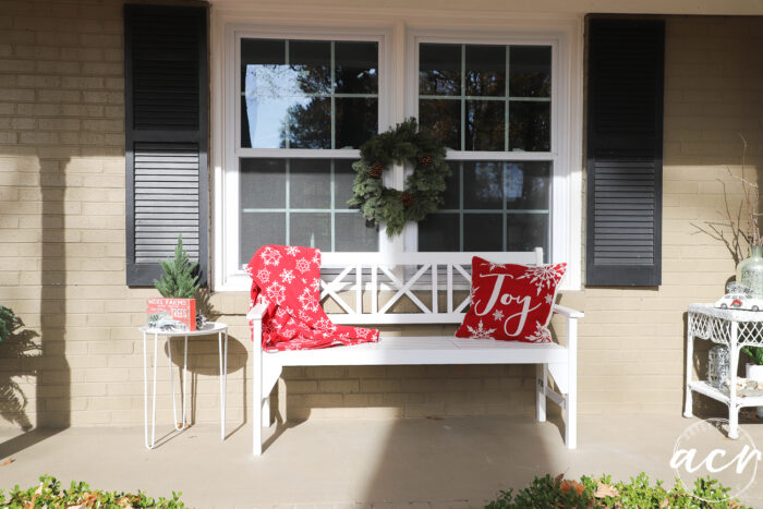 white bench on porch with red blanket green wreath on window