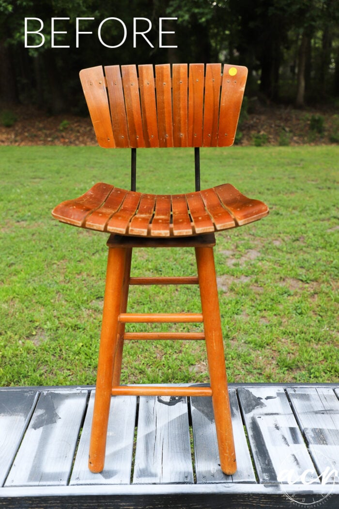 A tall mid century modern slatted bar stool with a curved back and seat stands outdoors on a painted wooden surface, with green grass and trees in the background. The word BEFORE is written at the top, hinting at an upcoming makeover.