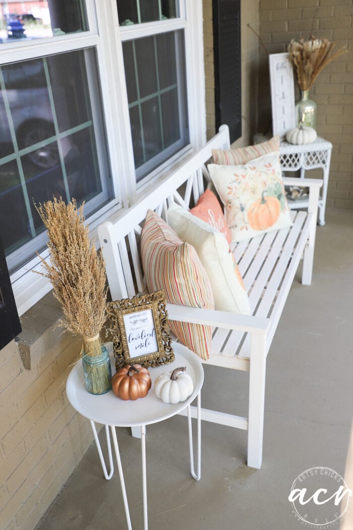 white bench with rust and orange colored pillows beside white table covered in fall items