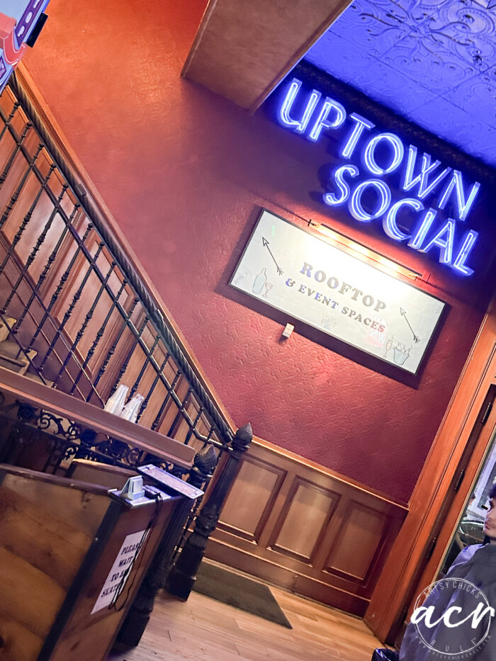 Staircase next to a red wall with a neon UPTOWN SOCIAL sign above and a ROOFTOP & EVENT SPACES sign below—one of the must-see things to do in downtown Charleston; part of a wooden railing and counter are visible in the foreground.