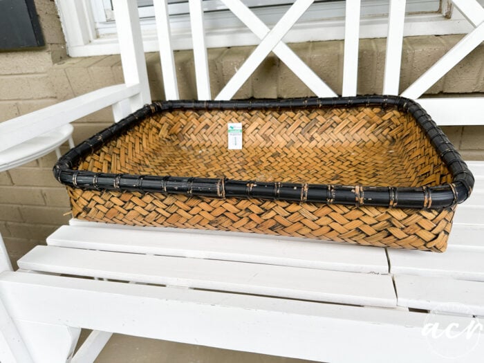 A rectangular painted flat basket with a black rim sits on a white wooden bench outdoors. There is a small price tag attached to the inside of the basket, offering an update on its value.