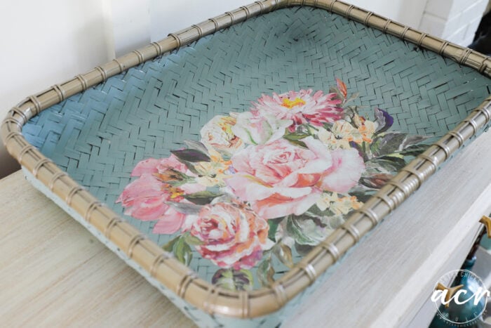 A rectangular painted flat basket tray in light blue with a metallic gold rim, featuring a vibrant floral design of pink and white flowers at the center. This stylish update rests elegantly on a light-colored surface.