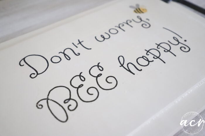 A white sign with whimsical black lettering reads &ldquo;Don&rsquo;t worry, BEE happy!&rdquo; A small cartoon bee hovers above the word &ldquo;BEE,&rdquo; giving this piece a charming makeover with napkin art.