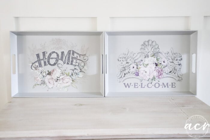 Two decorative trays on a light wooden surface. The left tray features the word HOME with floral designs, and the right has WELCOME in an ornate style. Ideal for those interested in how to paint melamine with a soft, vintage touch. Both include handles for easy serving.