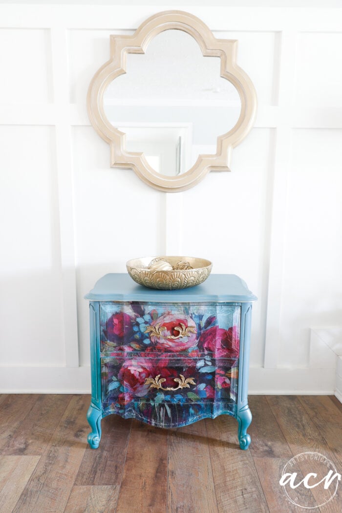 blue nightstand with colorful front, gold bowl on top, white wall and gold mirror above