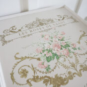 A decorative sign with gold ornate borders, pink roses, and green leaves evokes the elegance of a vintage french country style side table. The text Les Roses de Saadi tops this charming piece.