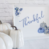 A blue pumpkin thankful tray, featuring "Thankful" in blue cursive with blue floral and pumpkin accents, is displayed beside three white pumpkins on a white surface against a white brick wall.