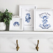 Create Inexpensive Artwork with Transfers A white mantle with gold hooks holds a potted plant and three framed blue and white prints, including one labeled Maison de Paris, showing how you can create inexpensive artwork with transfers against a light-colored wall.