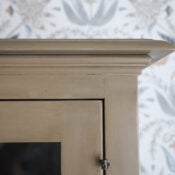 Close-up of the top corner of a beige wooden cabinet with a glass door, featuring a wood look finish, set against a background of floral-patterned wallpaper in blue, gray, and beige tones.