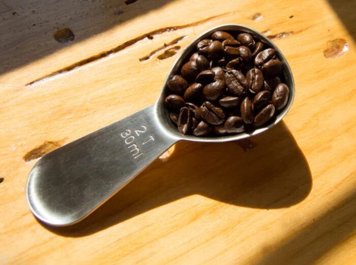 A metal tablespoon labeled 2 T 30 ml, an amazon finds favorite, holds whole coffee beans on a wooden surface in sunlight, casting a distinct shadow.