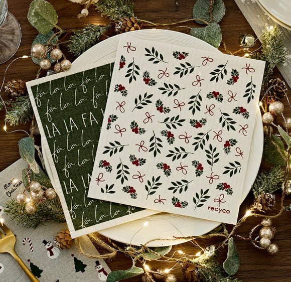 Two festive napkins on a plate: one with green leaves, red berries, and bows; the other with &ldquo;Fa La La La&rdquo; in white script on dark green. Decorative holiday lights and foliage surround this charming amazon finds table setting.