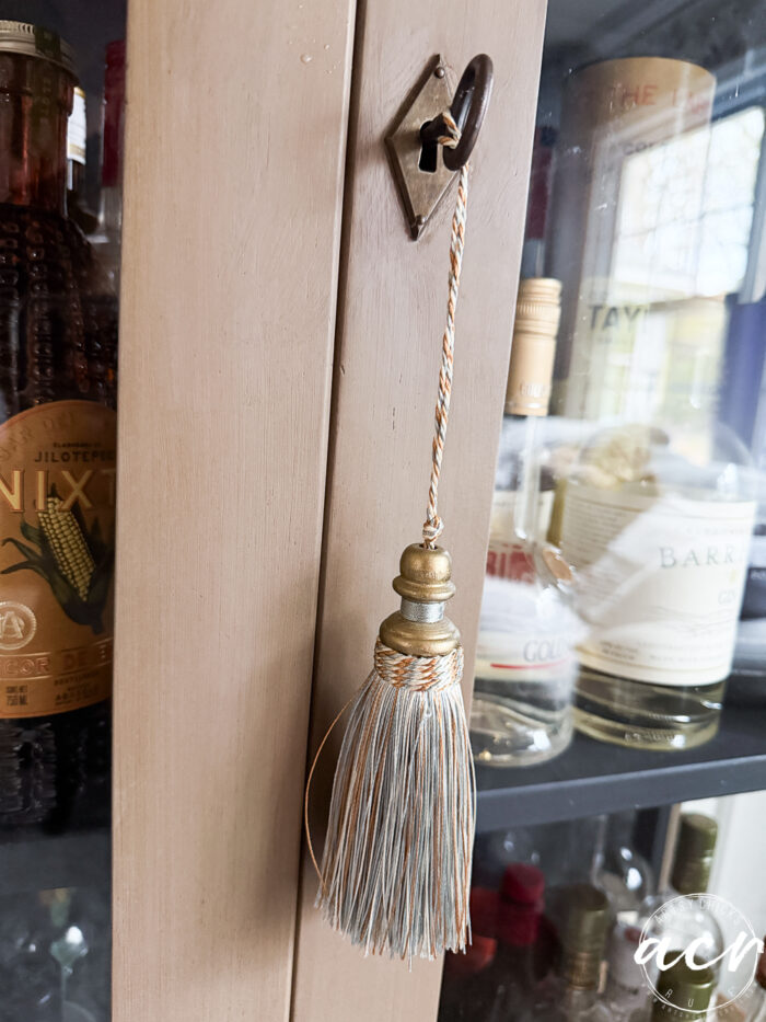 A gold and silver decorative tassel hangs from a diamond-shaped hook on a wooden cabinet door, with various bottles of liquor visible inside&mdash;one of those unique amazon finds that adds elegance to any home bar.