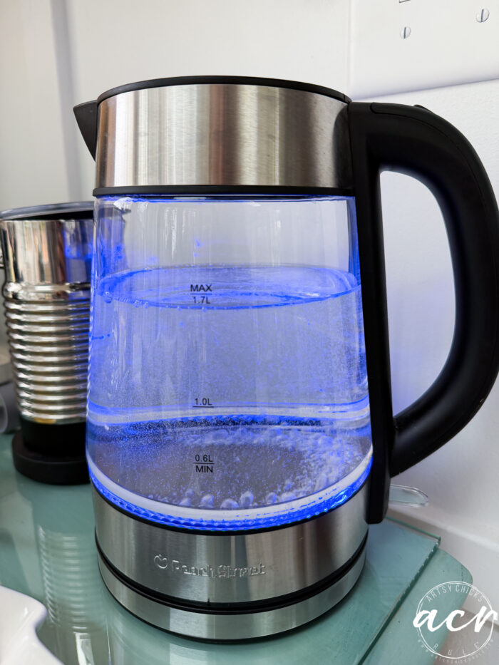 A glass electric kettle with stainless steel accents heats water, illuminated by a blue LED light&mdash;one of those amazon finds that elevates any kitchen. The water inside is starting to bubble, while a coffee pod machine sits in the background on the left.