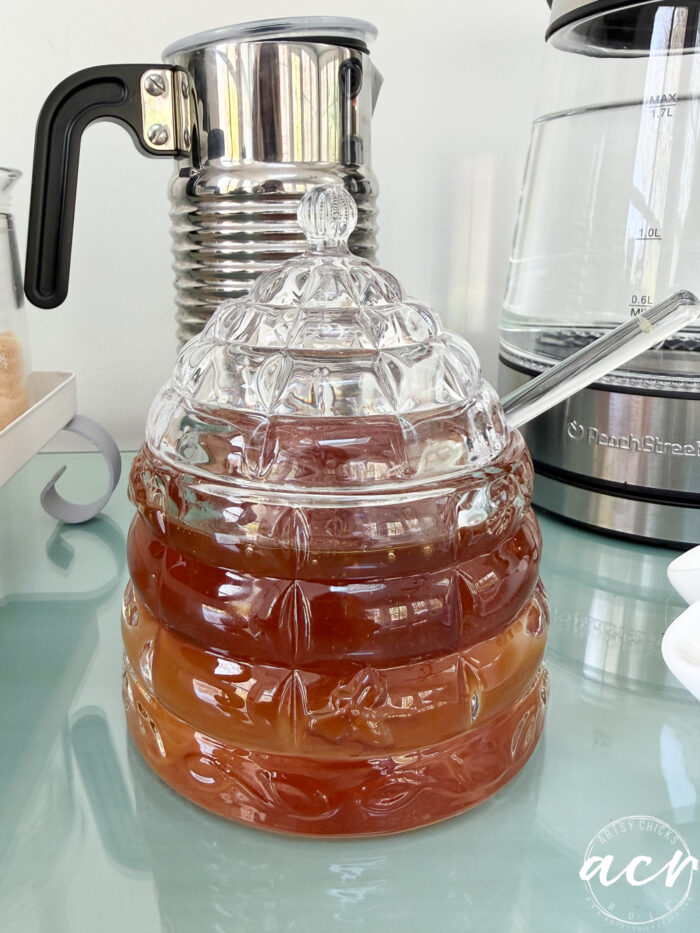 A glass container shaped like a beehive, filled with layered amber and honey-colored liquid, sits on a glass surface&mdash;a stunning amazon find. In the background are a metal frother and a glass electric kettle.