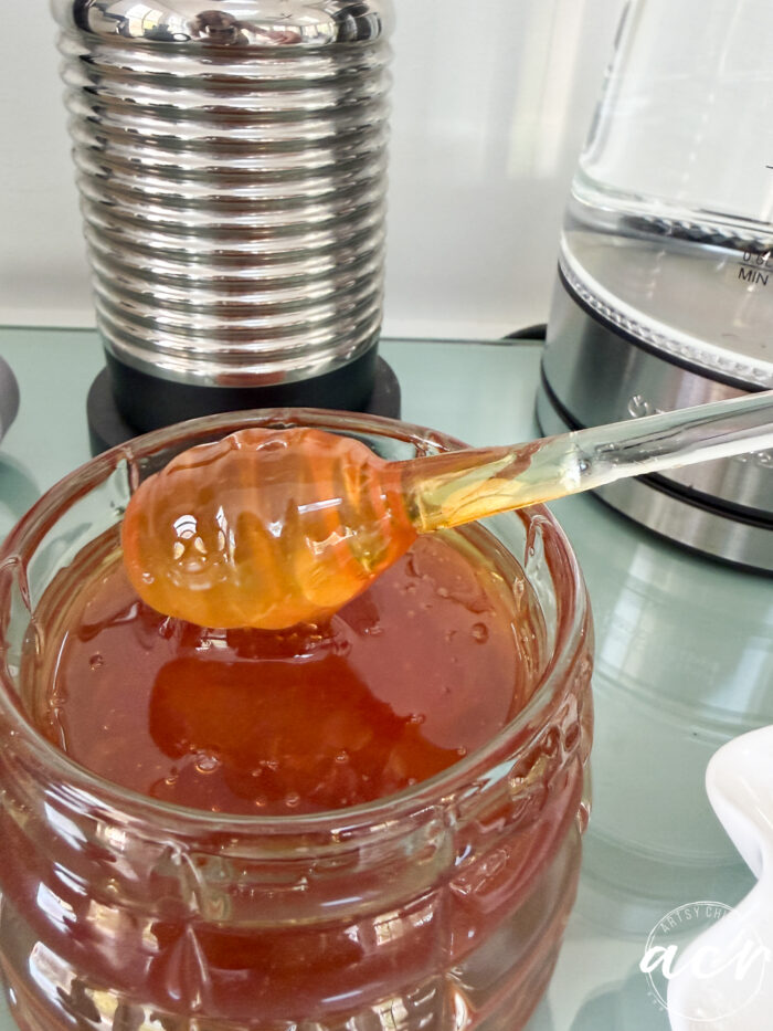 A glass honey dipper drips golden honey over an open jar, while a metal container and an electric kettle sit in the background&mdash;one of those amazon finds perfect for elevating your kitchen counter.