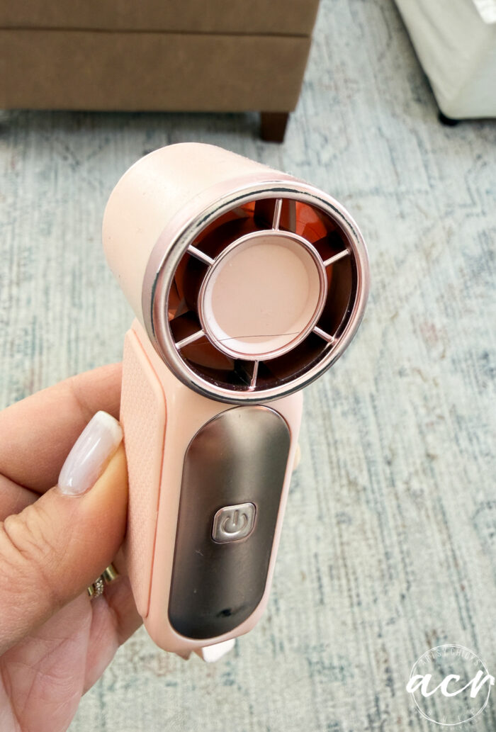 A hand holds a small, pink portable device with a circular vent and power button, resembling a mini handheld fan or dryer&mdash;one of those clever Amazon finds&mdash;against a background of a light-colored rug and furniture.