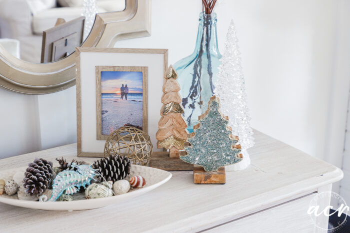 small dish with pine cones beside 3 trees, one of wood, one with green paint and one crystal clear one