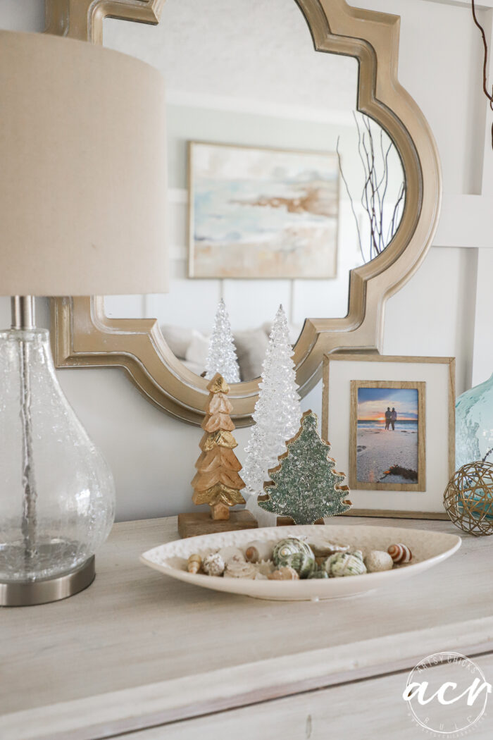 table top with dish of shells and 3 varying height trees behind it, one simple wood, one crystal white and one green sparkled.