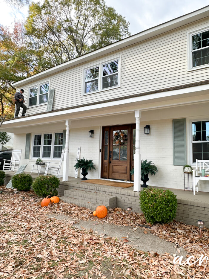 off white home with white trim and wood front door, man on roof attaching shutters