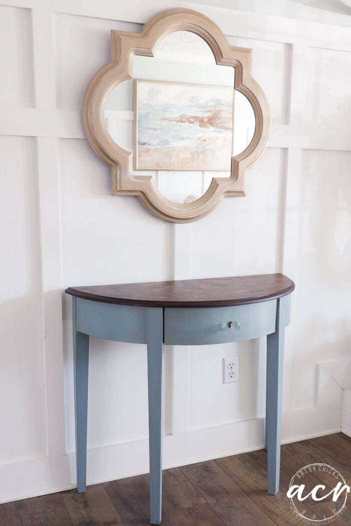 white wall with gold mirror and blue half moon table with stained top and crystal knob