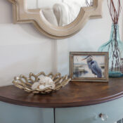 A small half moon table makeover with paint and stain features a wooden top, holding a decorative metal bowl with seashells, a framed bird photo, and a blue glass vase with twigs. Above the table, a mirror in a beige frame completes the look.