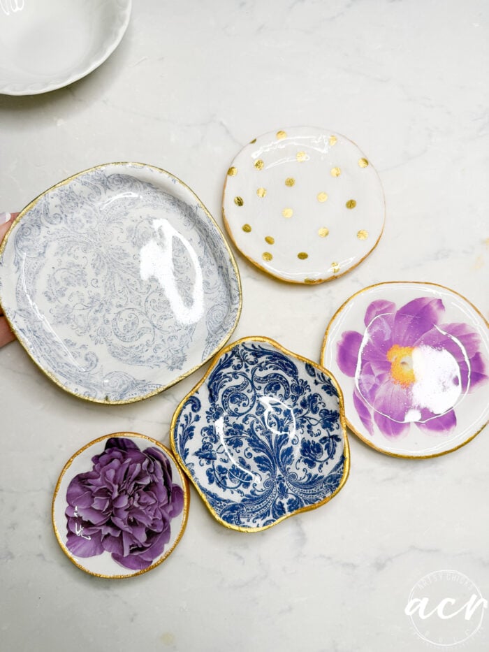 Five small, decorative ceramic plates with gold edges&mdash;perfect for diy air dry clay projects&mdash;are arranged on a white surface. Designs include blue patterns, gold polka dots, and purple flowers. A hand holds one of the plates.