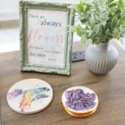 DIY Air Dry Clay (recipe and tips!) A wooden desk with a framed quote, a potted plant, a coaster with a colorful bird illustration, and a small dish holding purple flowers crafted from diy air dry clay. The corner features a notepad and acy watermark.
