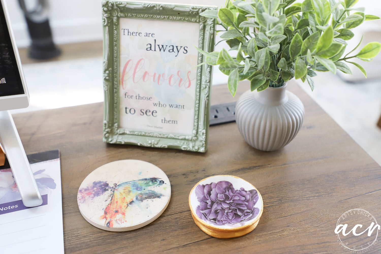 A wooden desk with a framed quote, a potted plant, a coaster with a colorful bird illustration, and a small dish holding purple flowers crafted from diy air dry clay. The corner features a notepad and acy watermark.