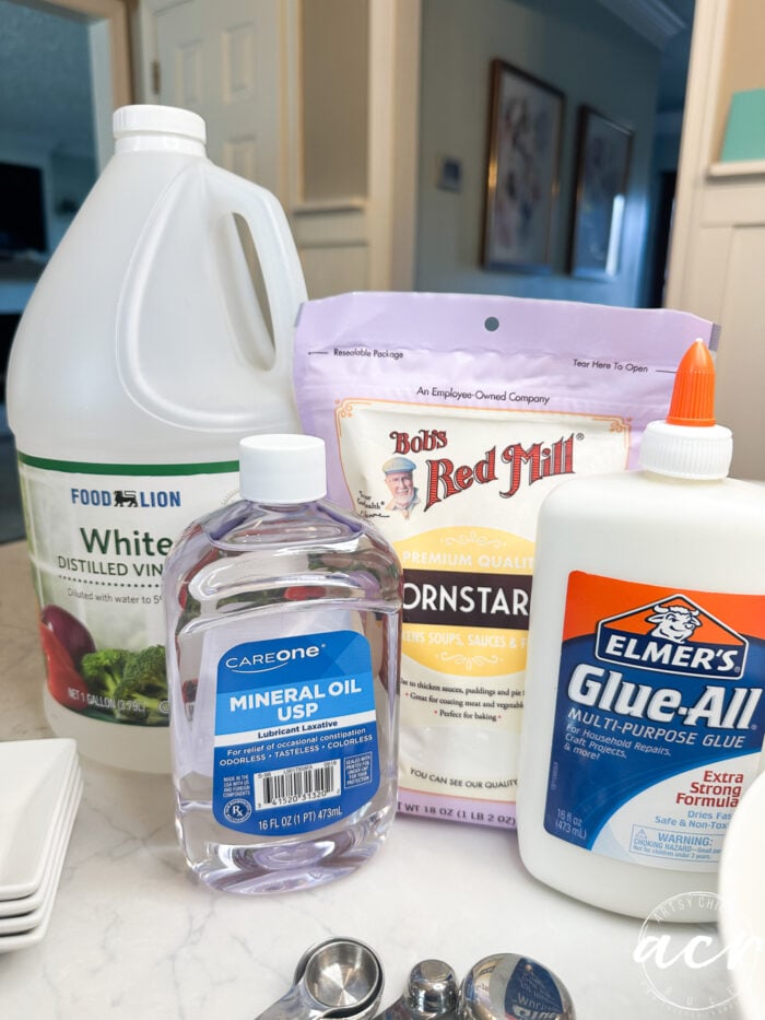 A collection of ingredients for diy air dry clay sits on a kitchen counter, including white distilled vinegar, cornstarch, mineral oil, and Elmer’s glue, with measuring spoons in the foreground.