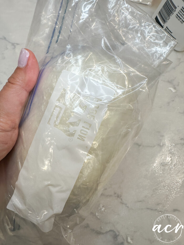 A hand holds a sealed plastic bag containing a white package of diy air dry clay. The clear bag rests on a light-colored countertop sprinkled with some scattered white residue.