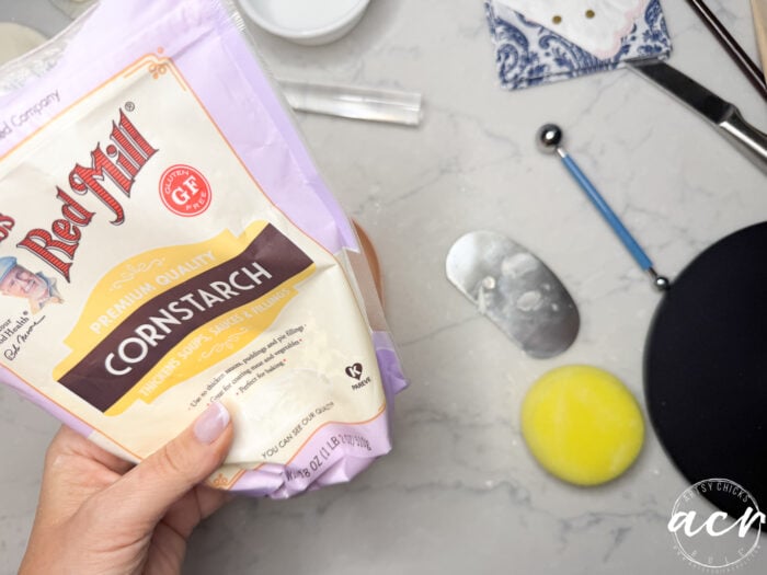 A hand holding a bag of Bob’s Red Mill cornstarch over a marble countertop, surrounded by tools like a metal scraper, melon baller, yellow sponge, and blue napkin—perfect for making diy air dry clay at home.