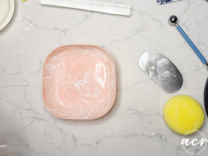 A square pink bowl dusted with flour sits on a marble countertop surrounded by baking tools, perfect for crafting diy air dry clay with a dough scraper, rolling pin, yellow sponge, and blue-handled utensil nearby.
