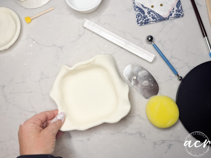 A hand holds an empty, fluted pie crust—perfect for a diy air dry clay project—on a marble surface, surrounded by baking tools like a yellow sponge, spatula, fondant roller, and a blue ball tool.